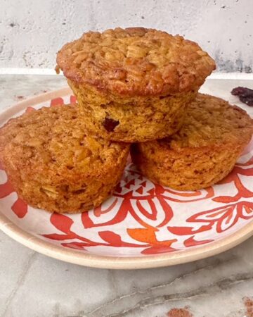 Stack of 3 pumpkin baked oatmeal cups on a white and orange plate with dried cranberries on the counter.