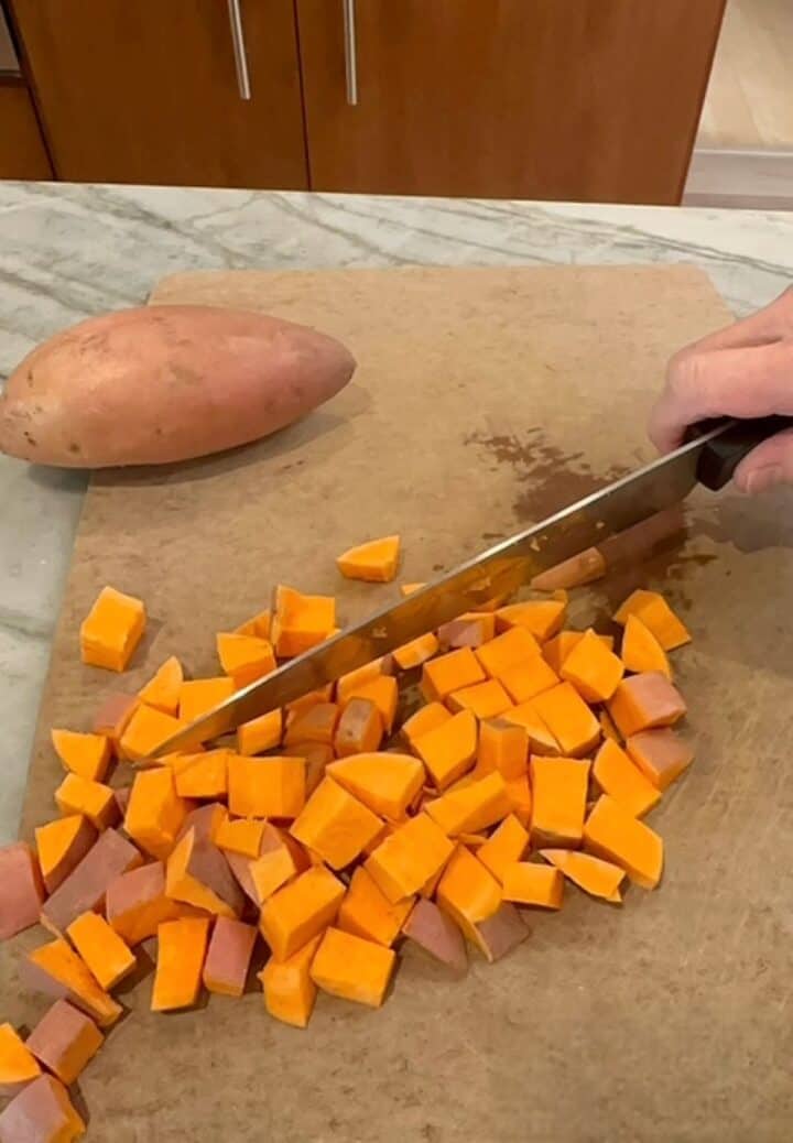 Orange sweet potatoes are cut into cubes by a large black-handled chef's knife on a brown cutting board.