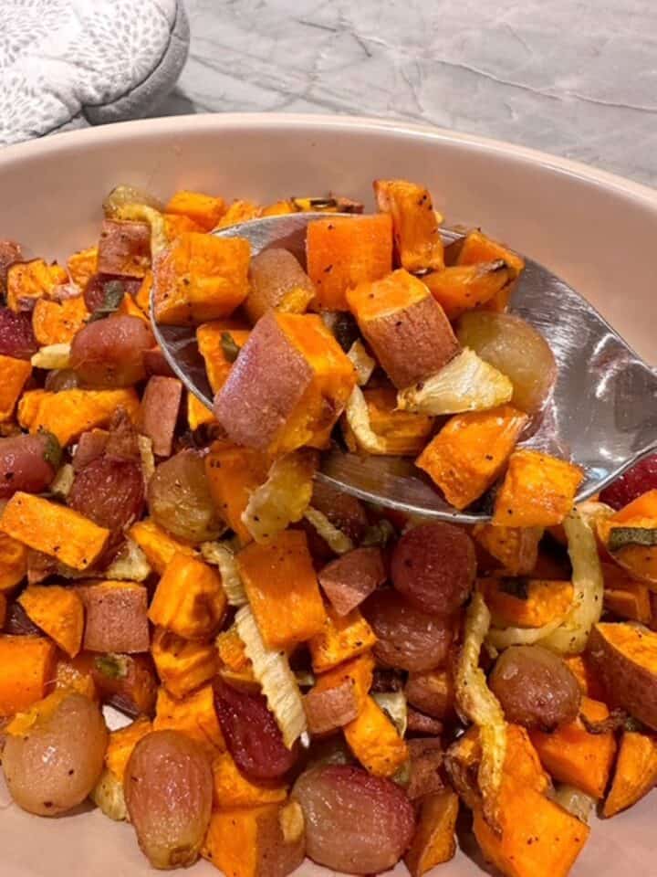 A big shiny spoon lifts roasted sweet potato bits and wrinkled grapes out of a round-edged casserole dish.