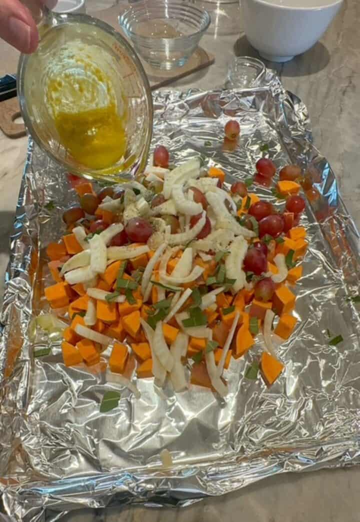 A golden-color sauce is poured over a mound of orange sweet potato cubes, white fennel slices, red grapes, and green sage on a shiny foil-lined pan.