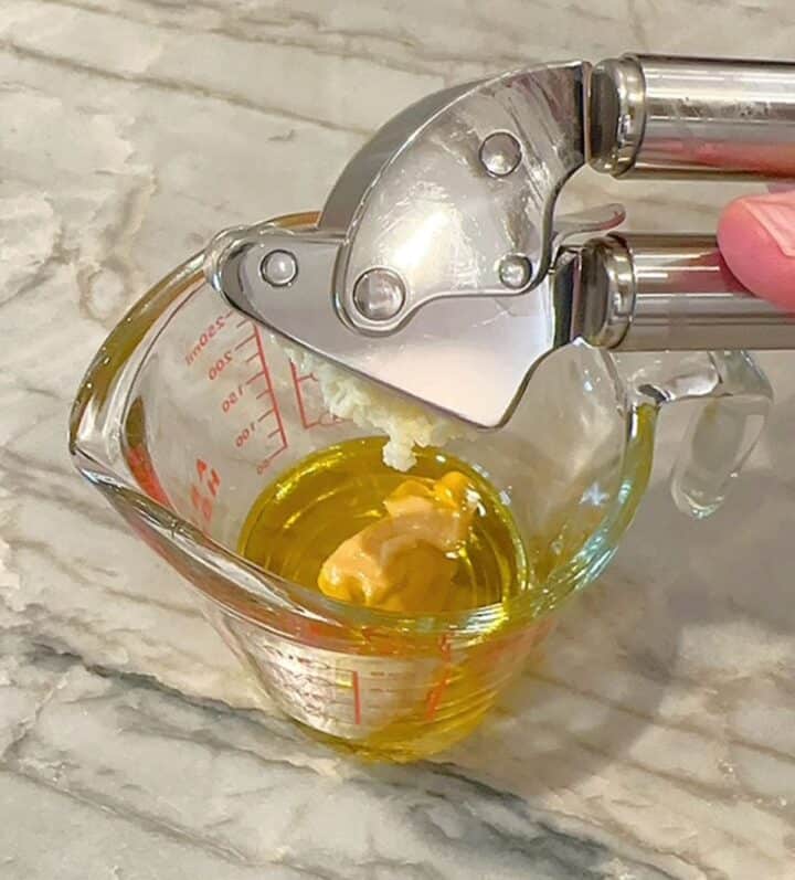 Shiny metal garlic press is squeezed over a small clear measuring cup with golden-green oil and mustard in it.