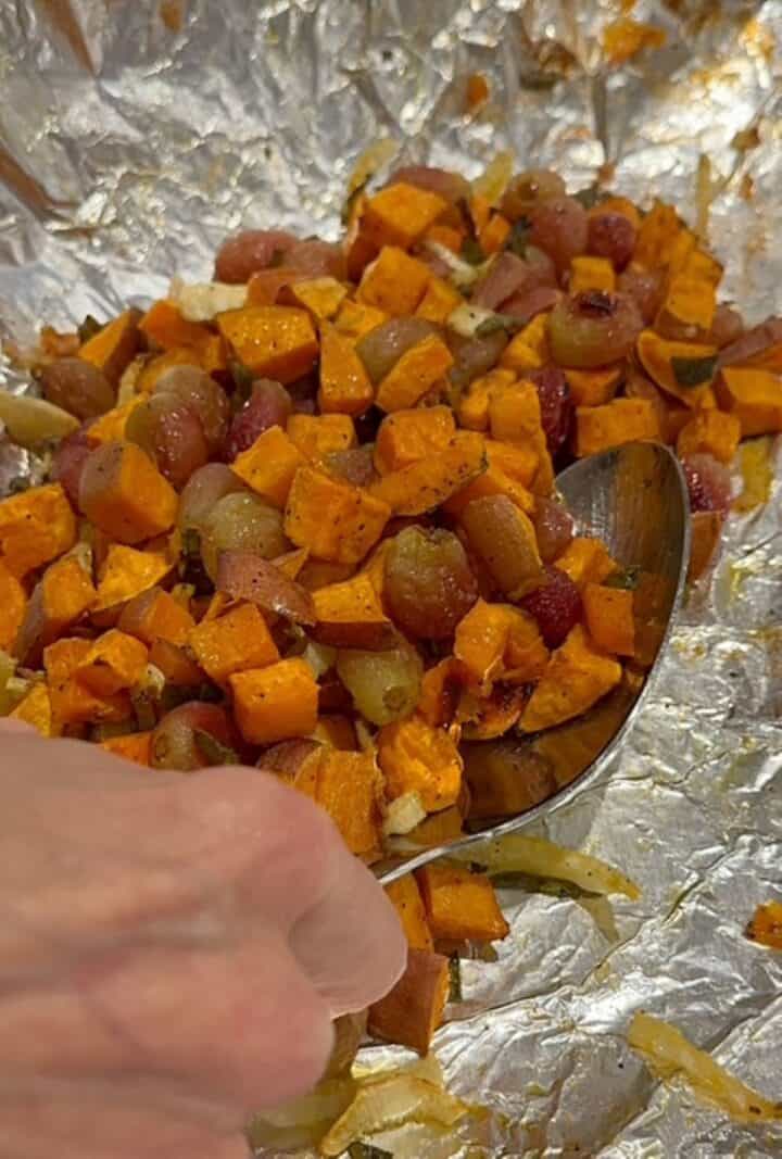 A large metal spoon scoops roasted orange sweet potatoes and wrinkled red grapes into a heap on a piece of foil.
