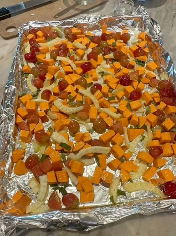 Orange sweet potato cubes, red grapes, white fennel slices spread out on shiny foil lining a sheet pan.