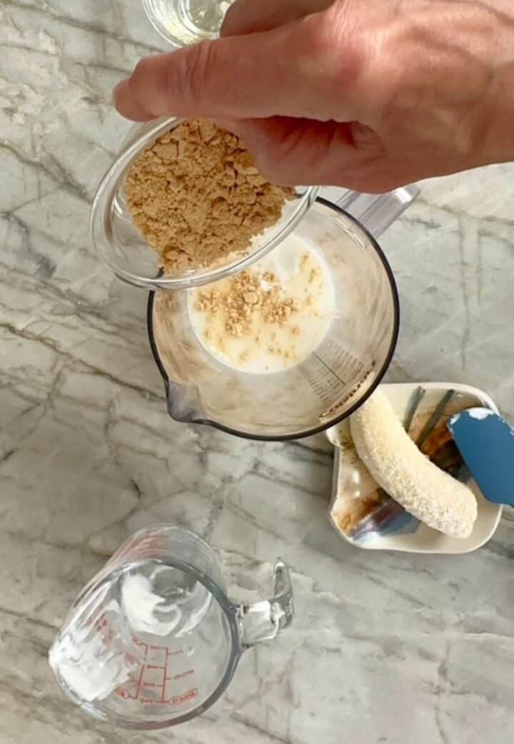 Pouring light brown powdered peanut butter into a clear blending container with white yogurt for a peanut butter smoothie.