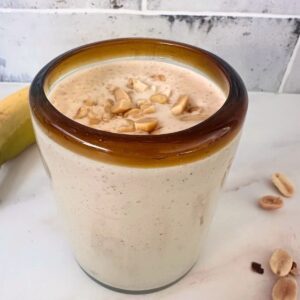 Clear glass with brown rim holds a creamy, pale brown peanut butter smoothie with chopped peanuts on top.