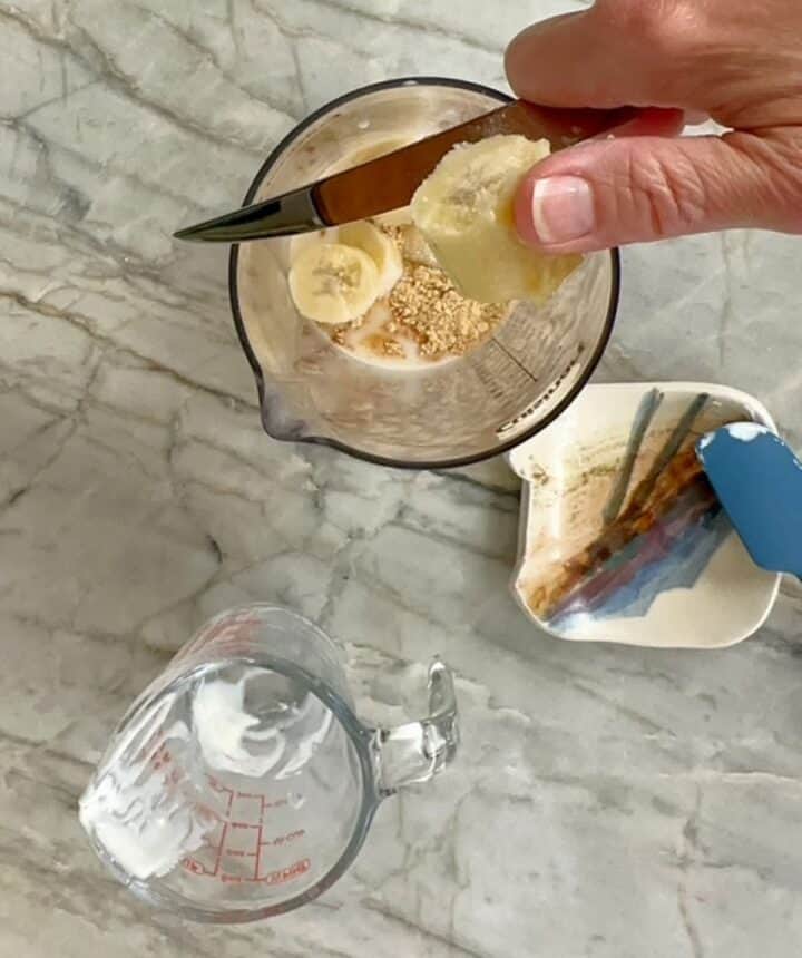 Knife slicing a banana into a clear blender container holding fixings for peanut butter smoothie.