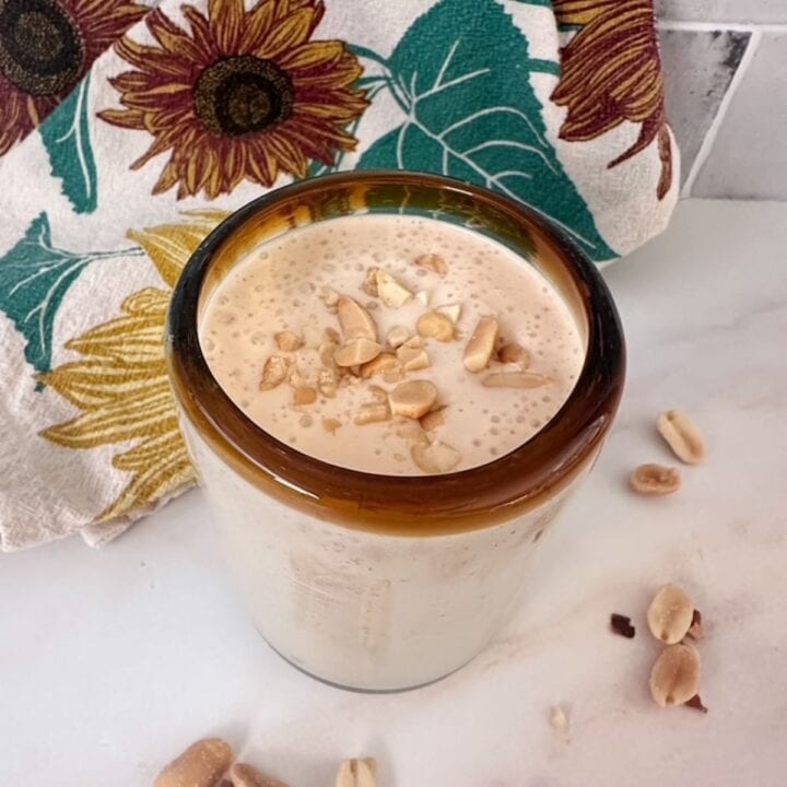 Clear glass filled with creamy, pale brown banana smoothie made with peanut butter powder and topped with chopped peanuts.