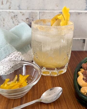 Short squat clear crystal glass full of yellow pear shrub and garnished with a sugar rim and slab of golden ginger.