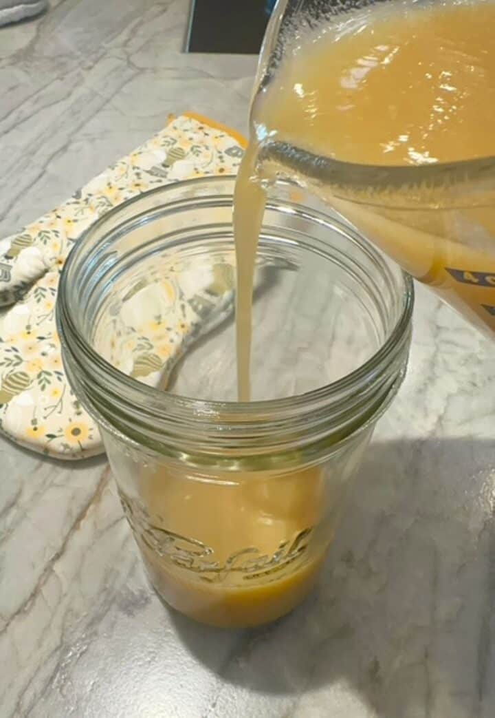 Amber pear-honey-ginger shrub concentrate is poured into a tall clear jar that has a lid for storage.