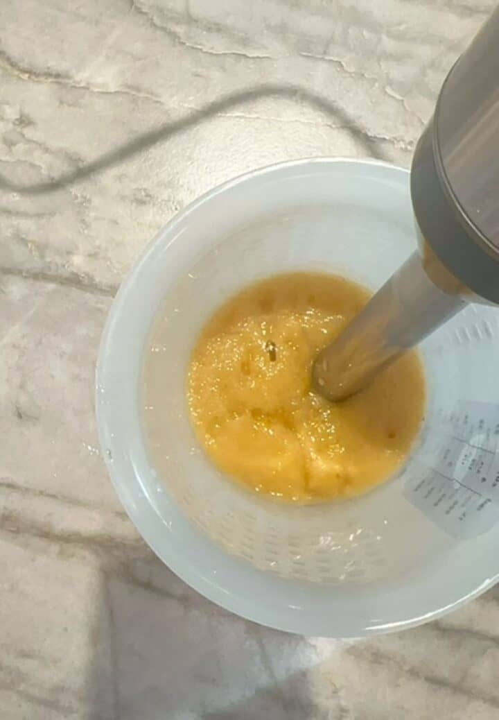Stick blender makes thick yellow puree out of soft pears in an opaque hi-sided container.