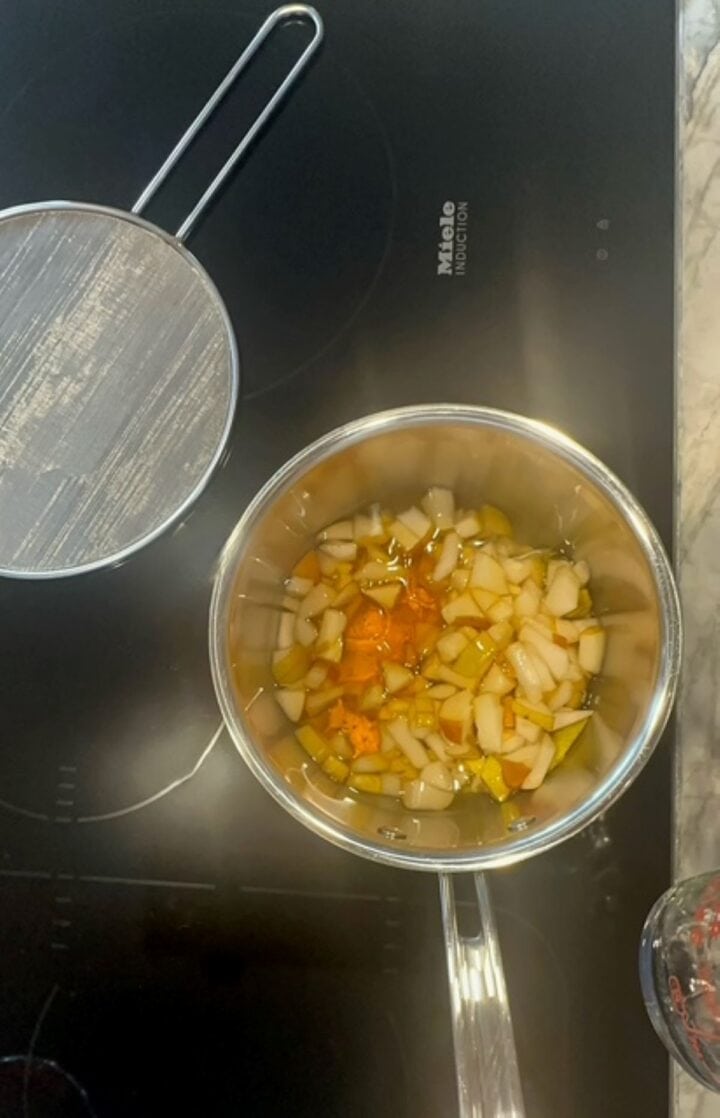 Metal saucepan filled with chopped pears and orange glob of honey sits on a black induction stovetop.