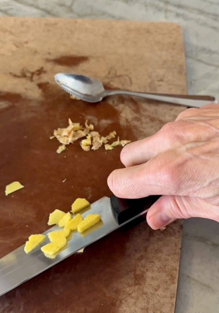 Slices of yellow ginger stick to a chef's knife on a brown cutting board.