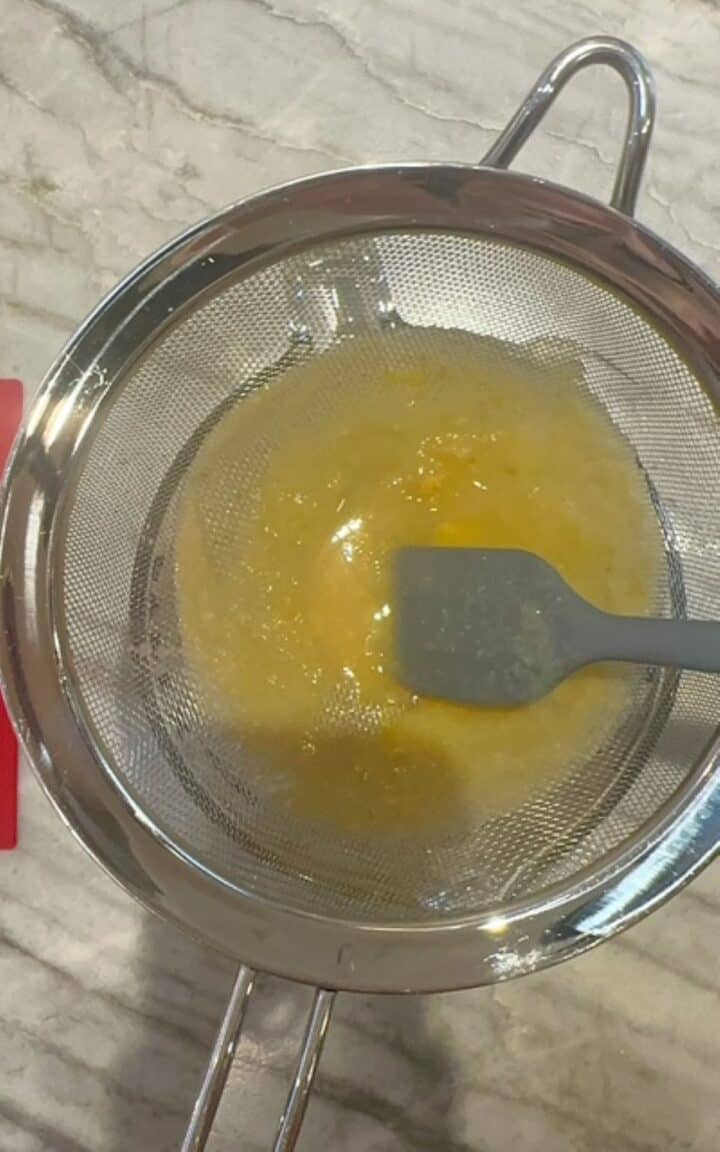 Gray silicone spatula pushes pureed pears through a strainer leaving a yellow ring behind in the strainer.