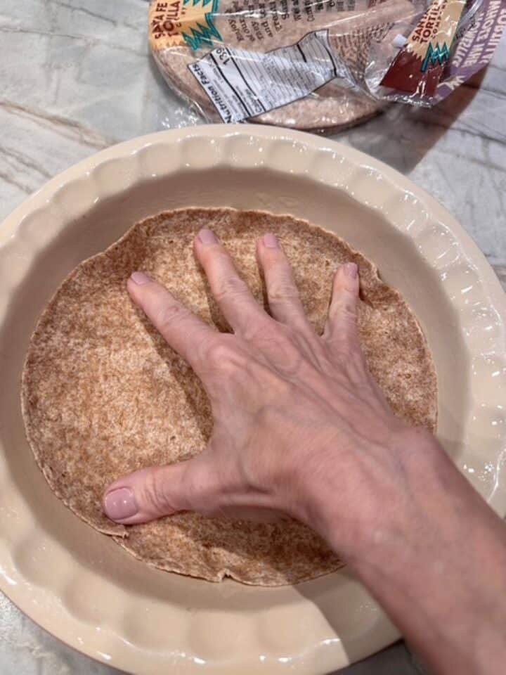 Hand presses a brown whole wheat tortilla into a beige ceramic pie dish.