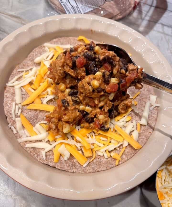 A mixture of taco-spiced ground meat, black beans, yellow corn is spooned over a brown wheat tortilla topped with orange shredded cheese.