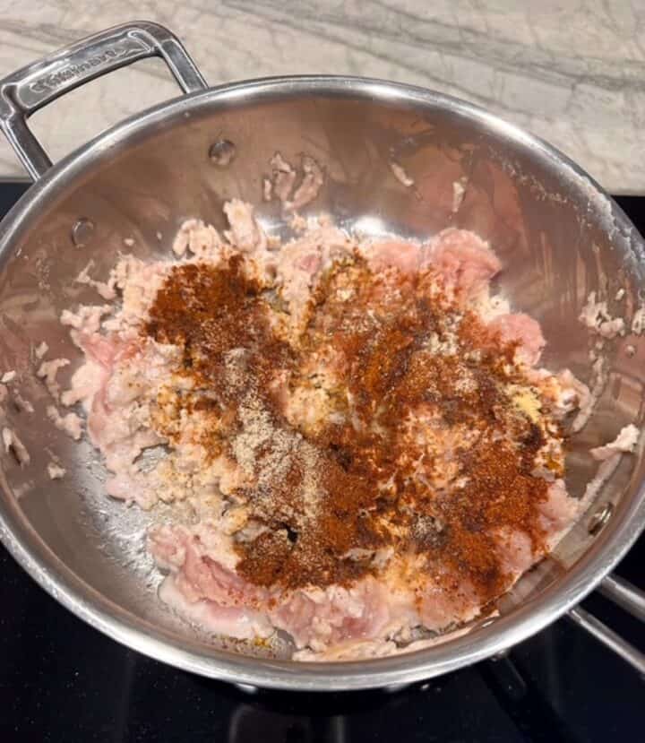Aluminum skillet with pink ground meat strewn with brown and red spices.