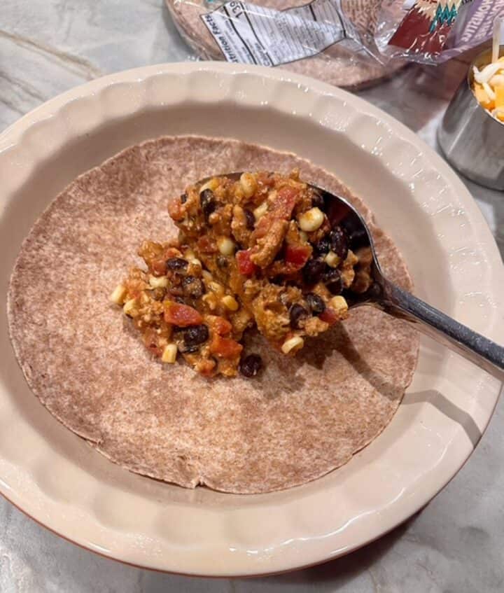 A mix of ground meat, tomatoes, black beans, and yellow corn is spooned over a brown wheat tortilla in a beige pie dish.