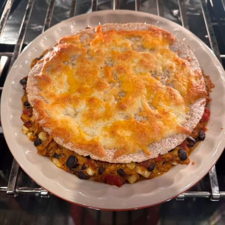 A pie dish holding a meat and bean taco filling is crowned with a tortilla covered in melted, crusty orange cheese.