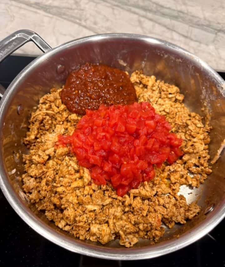 Red tomatoes and dark red salsa dollops on top of browned meat in a silver metal skillet.