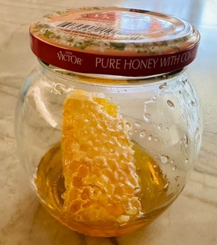Honeycomb dripping amber honey inside a rounded bottle with a brown lid.
