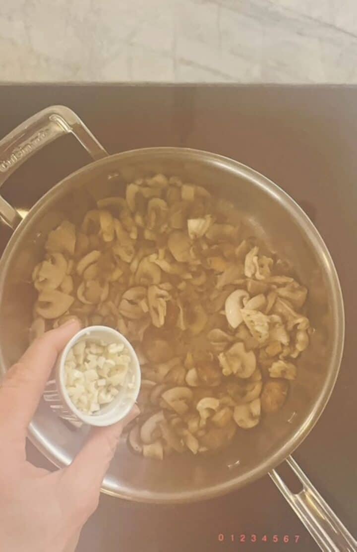 Pouring minced garlic into mushrooms cooking in large skillet for baked polenta sauce.