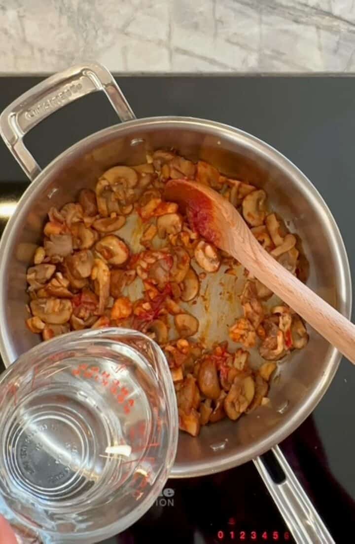 Pouring water into skillet with brown cooked mushrooms and red tomato paste to deglaze and cook sauce for baked polenta.