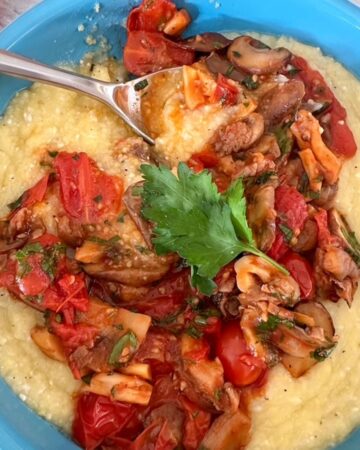 Round teal blue dish filled with creamy yellow baked polenta that has a red-brown topping of mushrooms and cherry tomatoes.