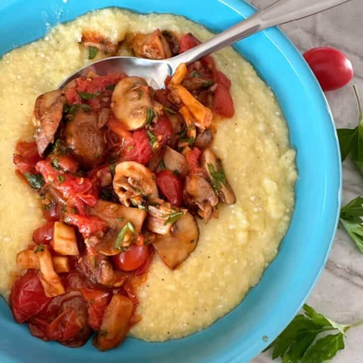 Round blue bowl hold creamy yellow baked polenta with a red-brown topping of saucy mushrooms and cherry tomatoes.
