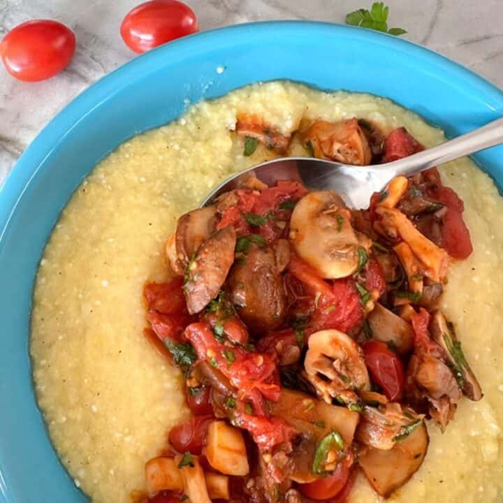 Creamy yellow baked polenta is topped with saucy cooked mushrooms and cherry tomatoes in this round, blue bowl.