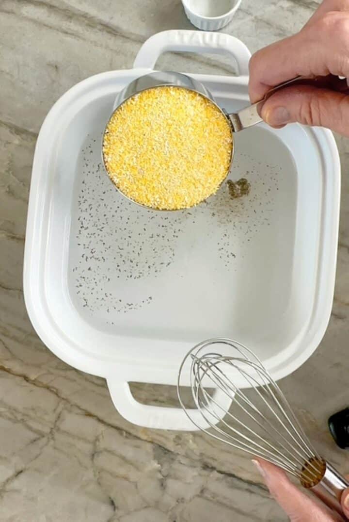 Pouring yellow polenta into water in a white casserole dish for baked polenta