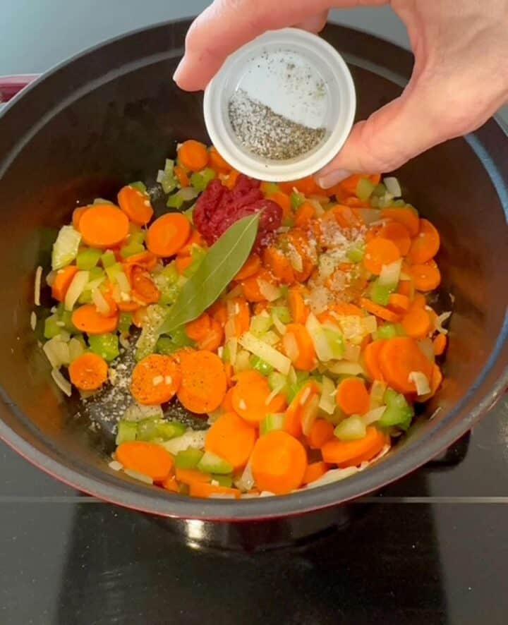 Salt and black pepper, dried green bay leaf, and red tomato paste are added to carrot-celery-onion veggies to add flavor to farro soup.