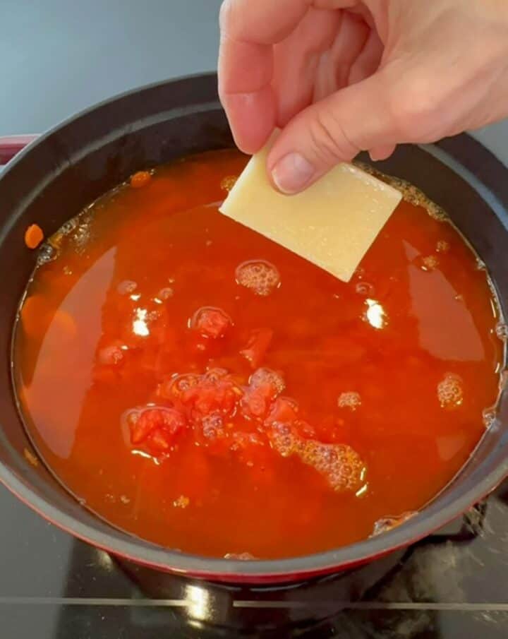 A pale yellow rectangular Parmesan rind is added to the brothy, red tomatoey looking farro soup.