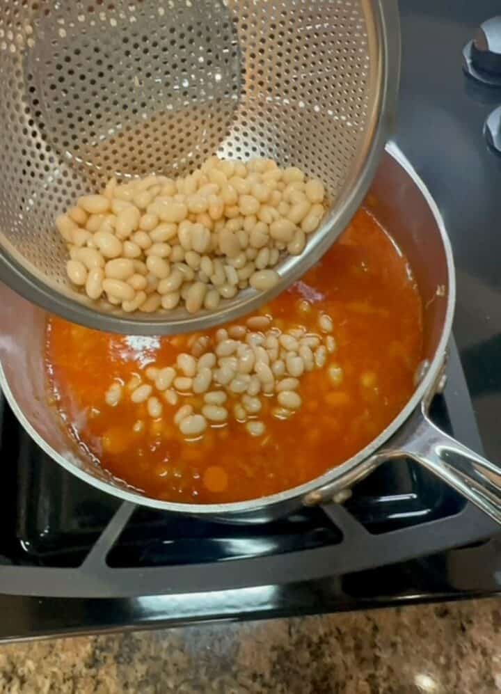 Pouring rinsed white beans into red-orange farro soup in a shiny pot.