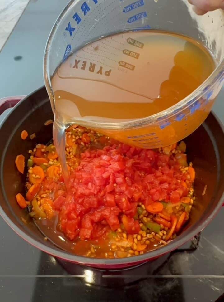 Golden colored vegetable broth is poured from a clear measuring cup into red pot that holds tomatoes, farro and vegetables for farro soup.