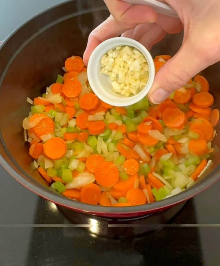 Creamy bits of chopped garlic are added to colorful farro soup vegetables from a white bowl.