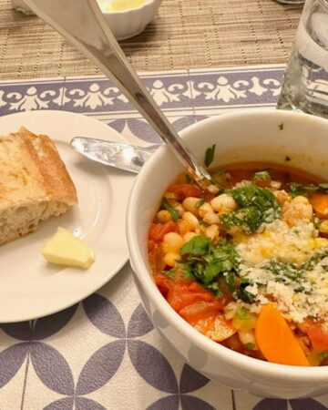 A golden piece of crusty bread and hearty white bean and farro soup with orange carrots sit on a patterned blue and white place mat ready for dinner.