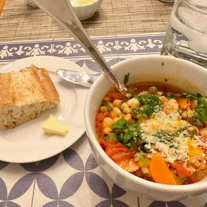 A golden piece of crusty bread and hearty white bean and farro soup with orange carrots sit on a patterned blue and white place mat ready for dinner.