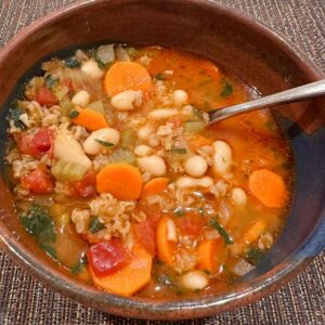 Brown pottery bowl filled with farro soup containing white beans, carrots, and tomatoes.