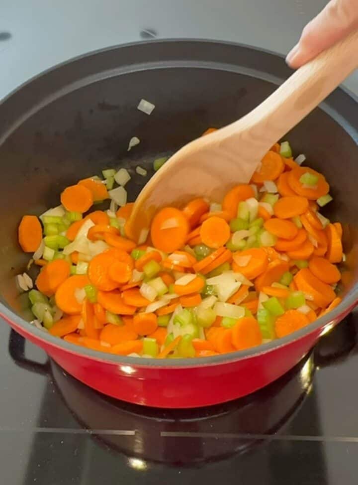 A brown wooden spoon stirs orange, white, and green carrots, celery, and onions softening in a red cocotte for farro soup.
