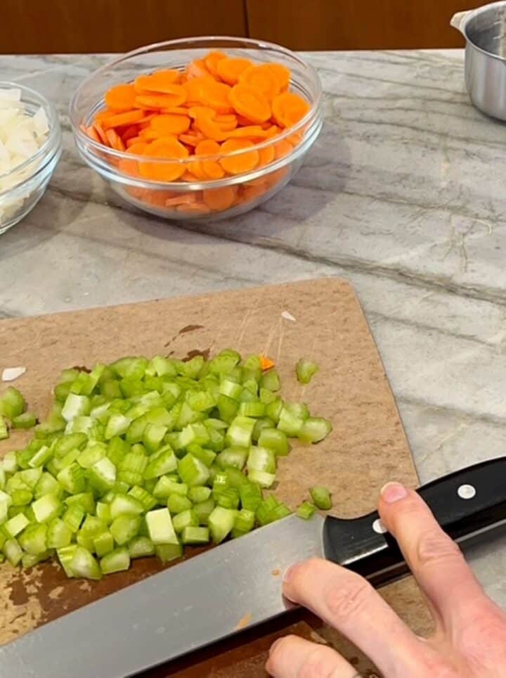 Diced green celery on a cutting board with chopped orange carrots and white onion nearby for the start of farro soup.
