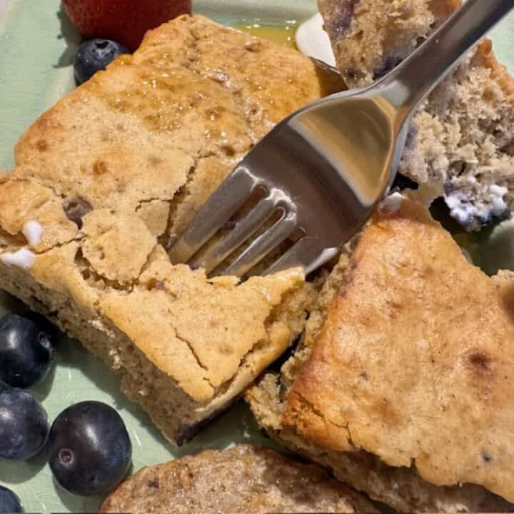 Fork digs into a fluffy golden rectangular blueberry pancake with berries next to it.