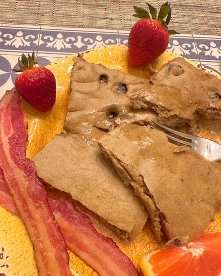 Square baked blueberry pancakes with strips of red bacon and red strawberries covered in syrup on a yellow plate.