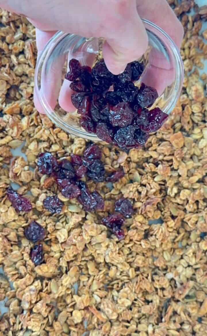 Dark dried cherries are sprinkled over golden baked granola in a sheet pan.