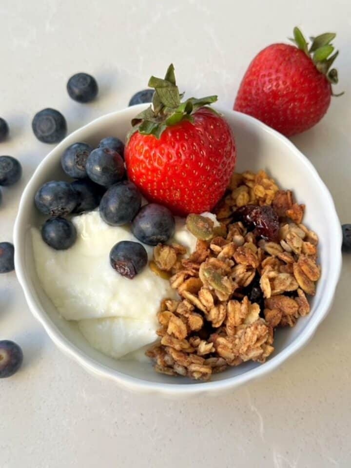 Blueberries and a whole strawberry plus golden oat and seed-rich granola top white plain yogurt in a white bowl.