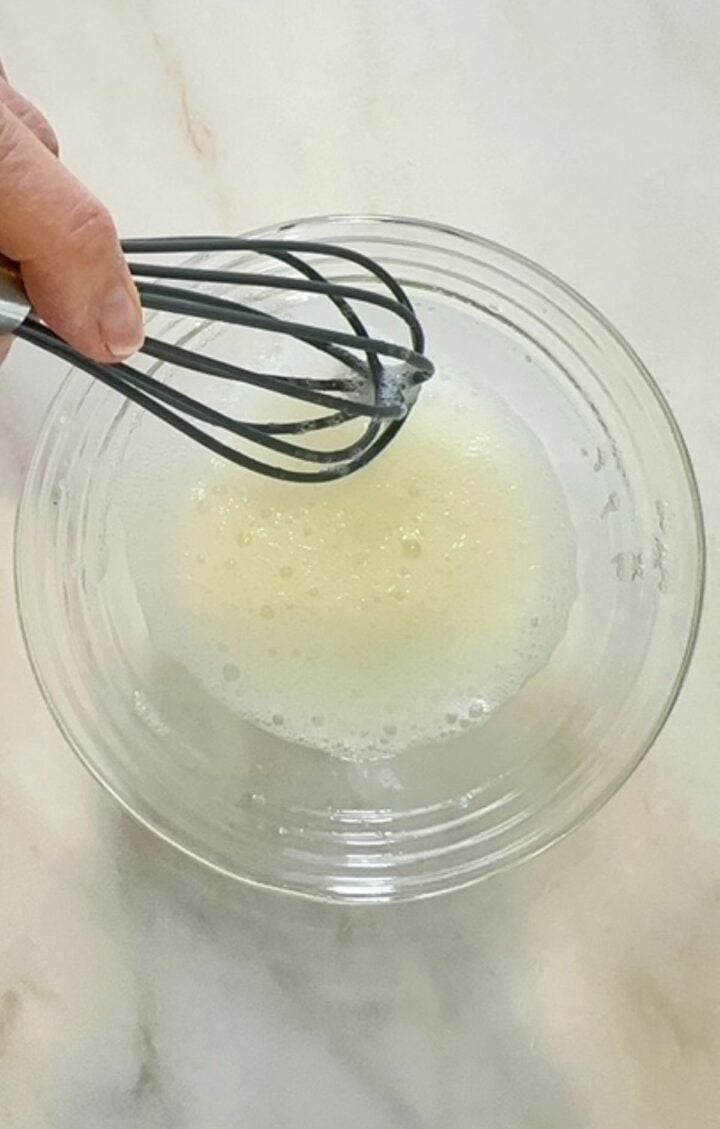 A whisk hovers over a clear bowl holding a frothy egg white.