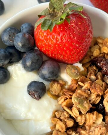 Closeup of white bowl with a red strawberry, blueberries, and golden granola over a plain white yogurt layer.