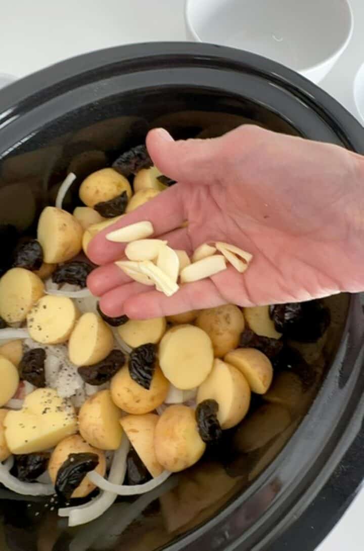 Creamy colored garlic cloves chopped in half are added to a slow cooker with chicken, prunes, and yellow potatoes.