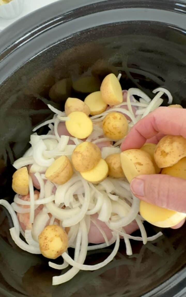 Yellow-gold baby potatoes are layered on top of white onions and chicken thighs in a black slow cooker liner.