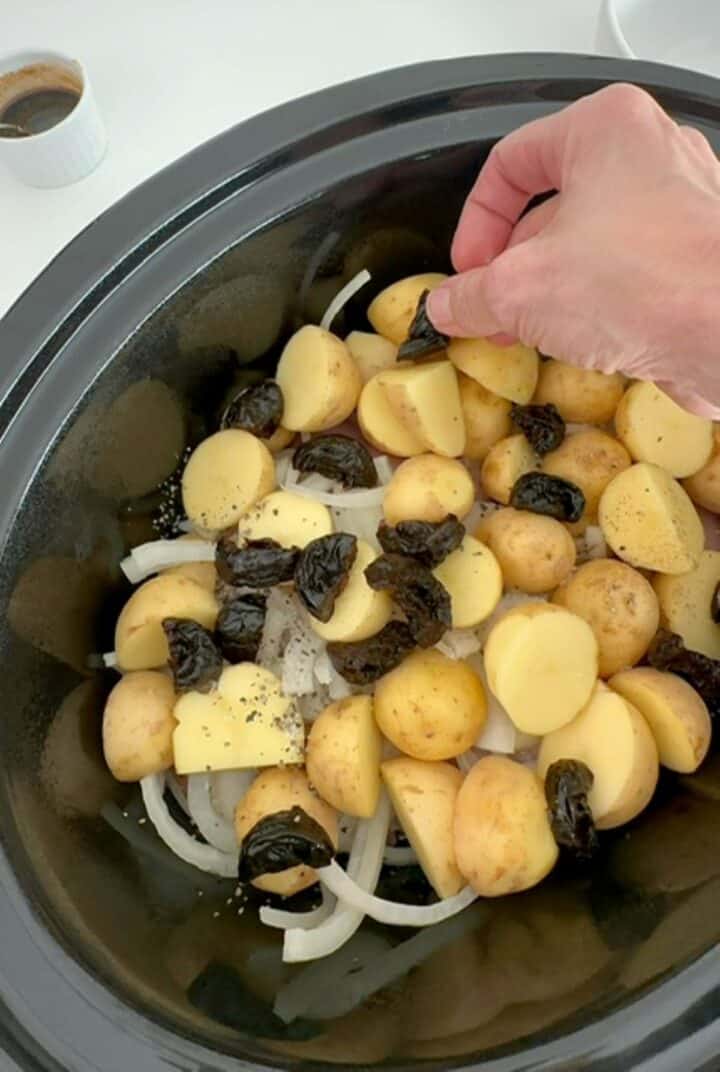 Dark black prunes are placed among potatoes and onions above chicken in a slow cooker.