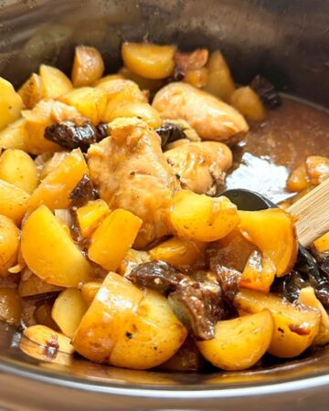 Cooked boneless chicken, yellow potatoes, and dark prunes are bathed in juices in a black slow cooker liner.